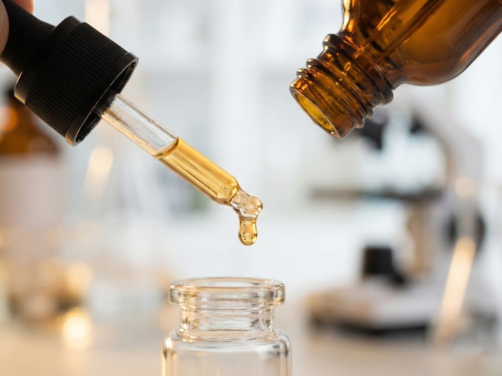 A professional macro shot of a precision glass dropper releasing a single drop of essential oil into a serum bottle in a laboratory setting, demonstrating XIMU’s titration accuracy and formula safety.